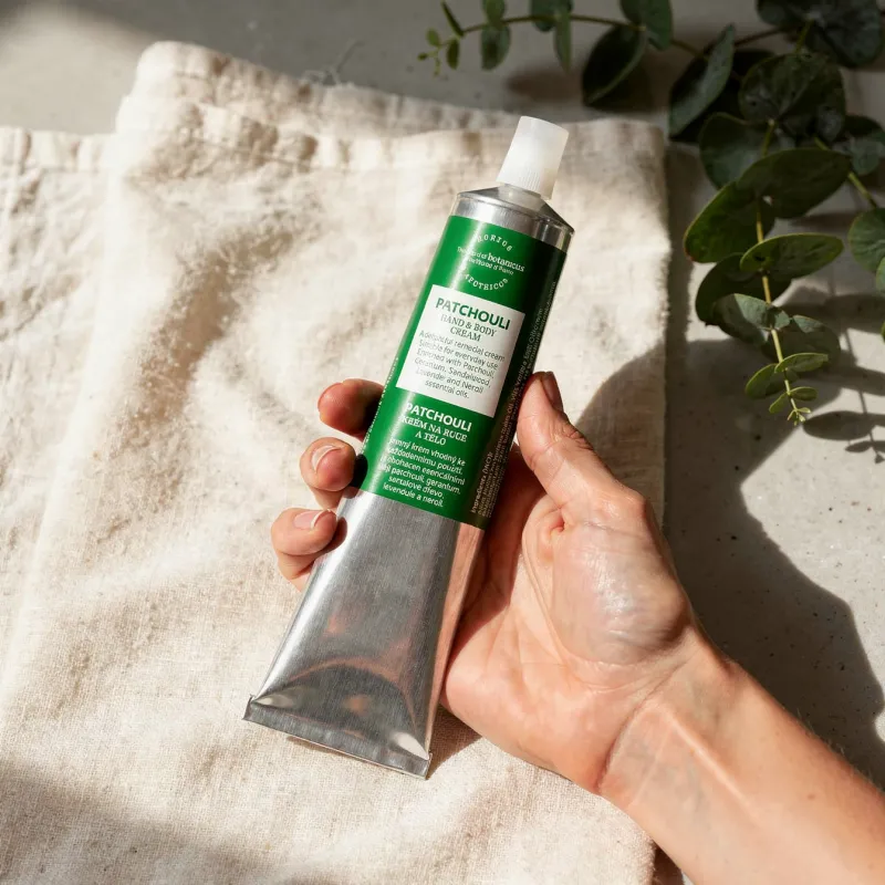 Tub crem patchouli în tub metalic, mâna ținând discul verde, pe o pânză bej, frunze în fundal.