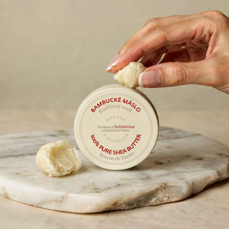 Mâna prelingând o bilă de unt de shea 100% pur, lângă cutia rotundă pe un bolovan de marmură, cu textul produsului pe etichetă.